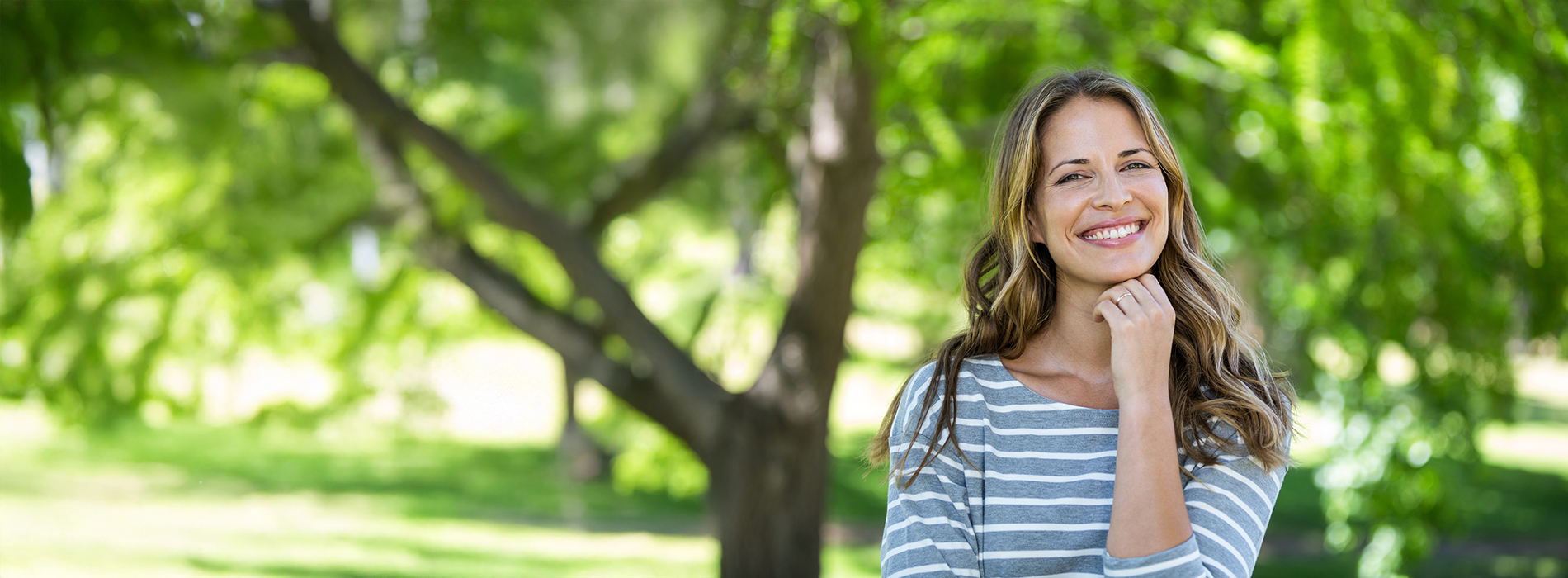The image shows a woman standing outdoors, smiling at the camera, with her hands placed on her hips.