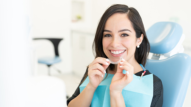 The image shows a woman seated at a dental chair, smiling at the camera while holding a toothbrush, with a dental office setting visible behind her.