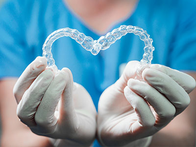 A dental professional s hands holding up a heart-shaped clear acrylic dental appliance against a blue background.