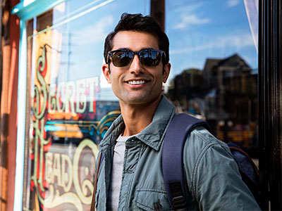 The image features a man standing outdoors, wearing sunglasses and a backpack, posing for a photograph with a cheerful expression in front of a storefront window with a sign that reads Sunday s.