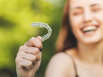 The image features a young woman holding up a clear dental retainer with her left hand.